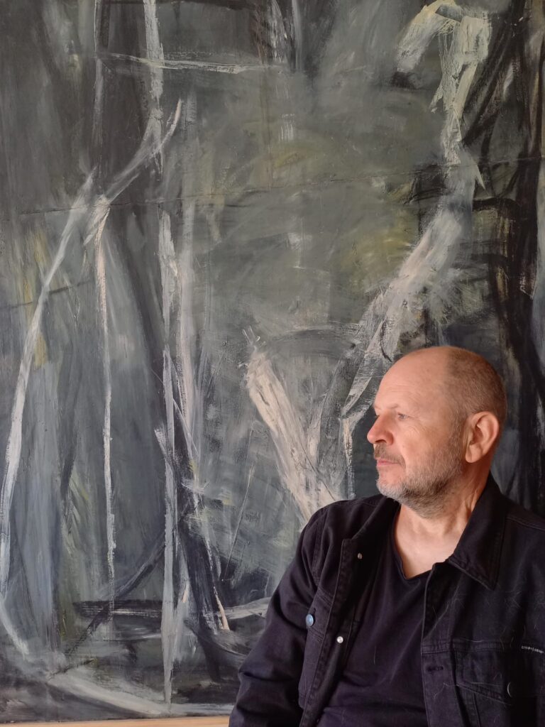 Artysta plastyk Roman Kasprowicz siedzi na tle dużego, abstrakcyjnego obrazu w odcieniach szarości i czerni, patrząc w bok; kompozycja podkreśla związek twórcy z jego malarstwem.