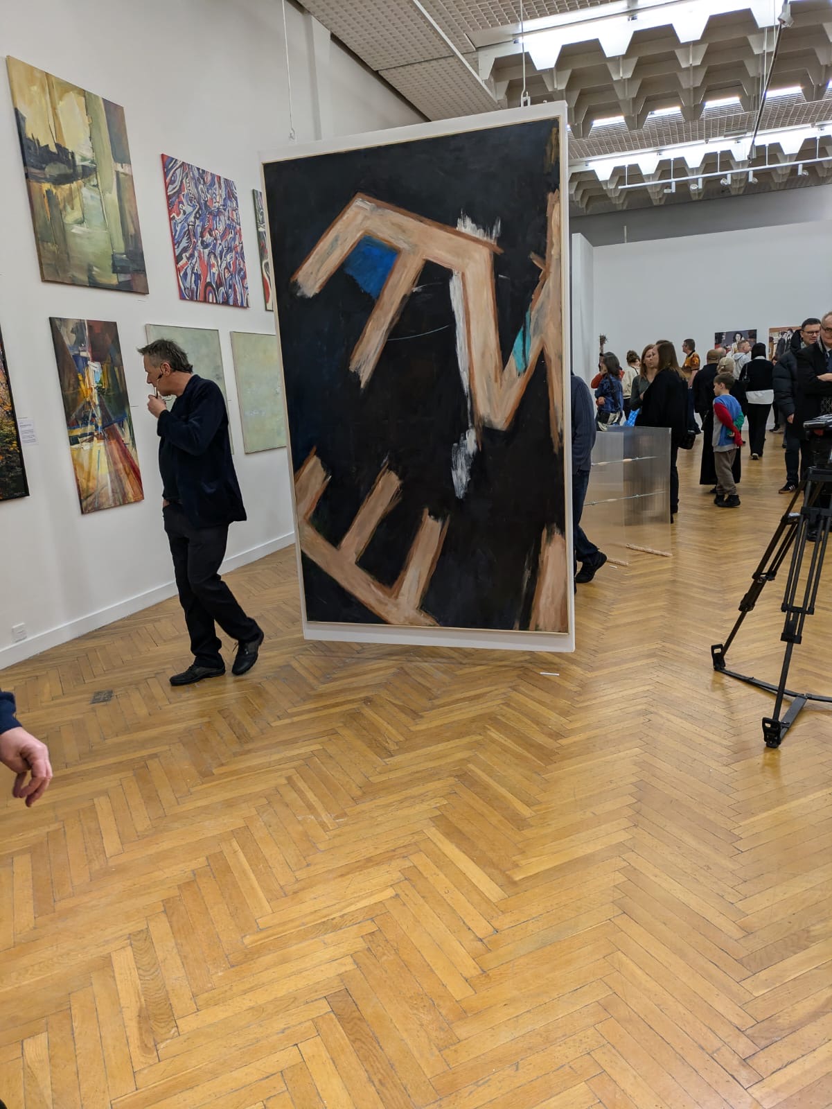 Wnętrze galerii sztuki z odwiedzającymi; na pierwszym planie duży, abstrakcyjny obraz olejny na płótnie zawieszony w przestrzeni, a na ścianach wokół inne prace malarskie oglądane przez gości.