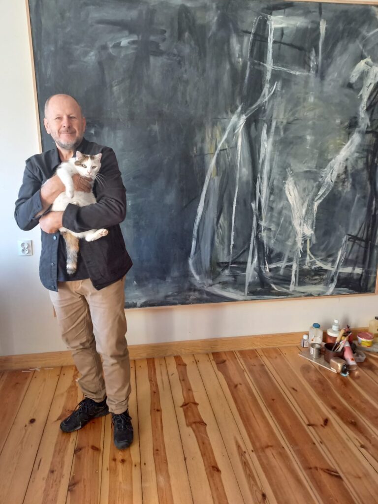Artysta plastyk Roman Kasprowicz stoi w pracowni na tle dużego, abstrakcyjnego obrazu, trzymając na rękach biało-rudego kota; na podłodze obok widoczne są farby i narzędzia malarskie.